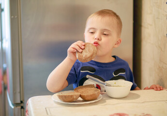 A little boy with a genetic disease Down Syndrome is sitting at the table and eating. The child himself holds a spoon. A special kid eats breakfast.
