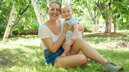 Portrait of happy smiling baby boy sitting with mother on grass under apple trees and looking in camera