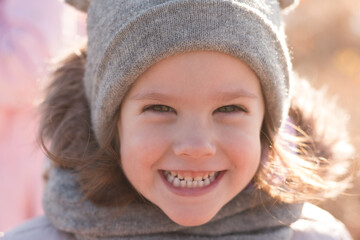 A small beautiful child with a wide smile in a funny hat, a fluffy hood, and a scarf holds a parent's hand on a walk outdoors