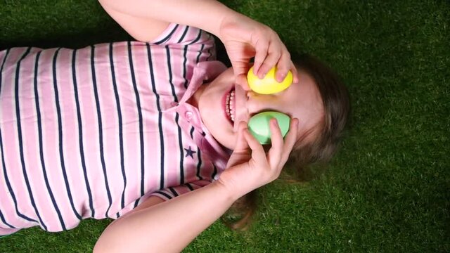 Little caucasian girl is having fun on the grass, closes her eyes with colored Easter eggs. Egg hunt, children's easter fun tradition