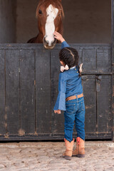 A girl in in cowgirl clothes touching a horse in a barn
