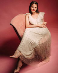 Smiling brunette girl in ivory flattering dress on salmon background. Gorgeous bridesmaid with pale...