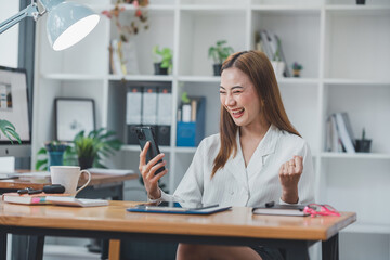 Obraz na płótnie Canvas Excited Asian woman sitting at her desk Feel satisfied with the business going well. Very happy to receive letters from laptops and smartphones promoted at work.