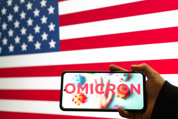 hand holding mobile phone with omicron COVID 19 variant strain in front of flag of USA showing news...