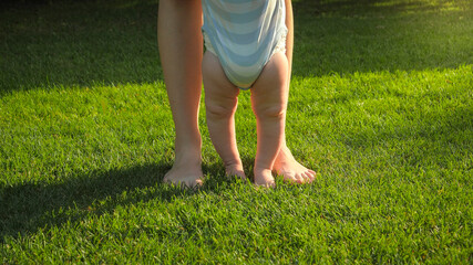 Closeup of barefoot baby boy and mother standing on fresh green grass at park. Concept of healthy lifestyle, child development and parenting.