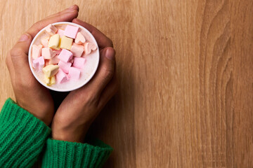 Top view of woman hands in green sweater holding a cup of coffee with pink and white marshmallows on the wooden background with lollipops. Christmas or New year concept        