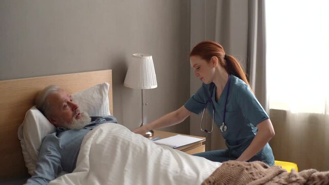 Smiling caring young female doctor helping senior adult male patient to lying on bed and covering him blanket at home. Cheerful nurse taking care of mature old patient and talking to him, slow motion.