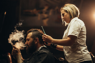 Young bearded man with a mohawk getting haircut by hairdresser and smoking a cigarette