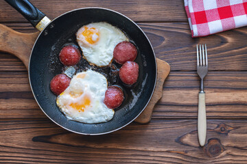 Fried eggs with sausage in a pan on a wooden table. View from above