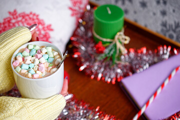 Close-up of a female hand in sweater with a mug with cocoa and marshmallow, for a cozy evening. Against the background of a wooden tray with a book, a candle, a pillow, tinsel and candy cane.