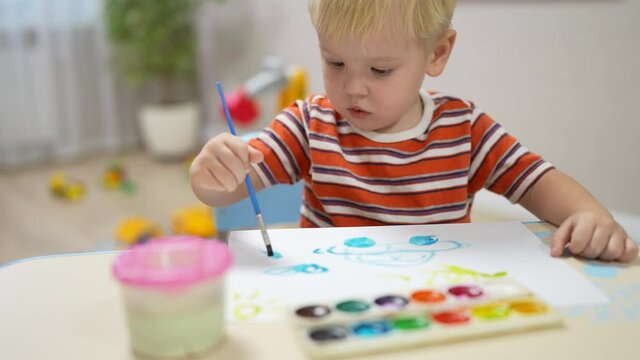 a little boy with blond hair sits at a table and draws a car with paints on paper.