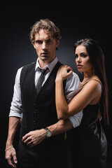 young sexy woman in slip dress hugging man in vest and shirt while looking at camera on black.