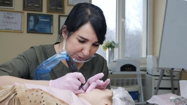 Beautician at work. Beautician wearing a protective mask applies a permanent pigment tattoo to women's lips with a tattoo needle in a salon