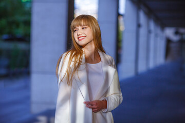 Happy young adult woman smiling with teeth smile, walking outdoors on city street at sunset time. Attractive girl weared in autumn clothes. White coat, long hair. Blurred details