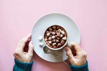 Mug with cocoa and marshmallows in female hands in knitted sweater on pink background. Top view,...