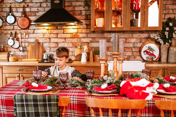 cute boy in knitted sweater and Santa hat in kitchen of chalet helps to set table for festive Christmas or New Year's dinner