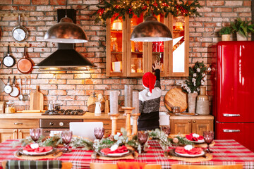 cute boy in knitted sweater and Santa hat in kitchen of chalet helps to set table for festive Christmas or New Year's dinner