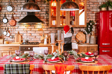 cute boy in knitted sweater and Santa hat in kitchen of chalet helps to set table for festive Christmas or New Year's dinner