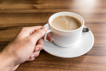 A man takes a cup of coffee on a saucer with his hand against the background of a wooden table in a cafe