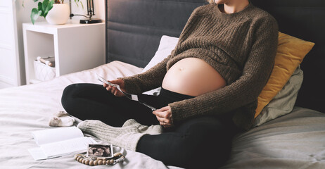 Pregnant woman with ultrasound image and medical test reports in cozy knitted sweater in bed at...