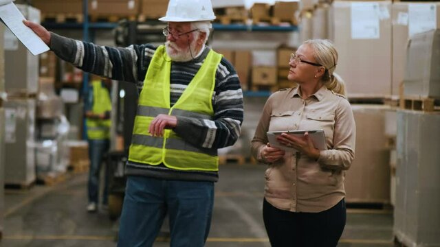 Female manager and old worker checking goods in warehouse using digital tablet. Industry and supply chain management concept. Slow motion