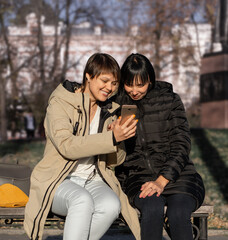Fototapeta na wymiar Two young women with a smile sit on a bench in the park and look into one smartphone. Selective focus