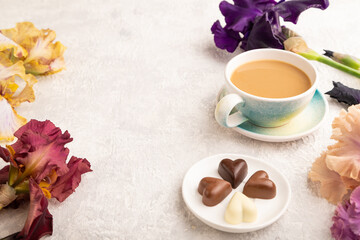 Cup of cioffee with chocolate candies and purple and burgundy iris flowers on gray concrete background. side view, copy space.
