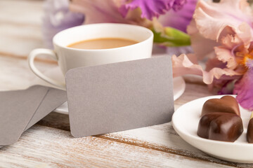 Gray business card with cup of cioffee, chocolate candies and iris flowers on white wooden background. side view, copy space.