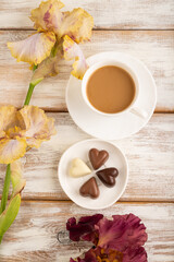 Cup of cioffee with chocolate candies, lilac and purple iris flowers on white wooden background. top view, close up.