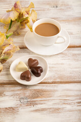 Cup of cioffee with chocolate candies, lilac and purple iris flowers on white wooden background. side view, close up.