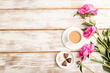 Cup of cioffee with chocolate candies, pink peony flowers on white wooden background. top view, copy space.
