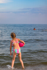 Fototapeta premium little girl, cheerful blonde, 6-7 years old, stands on the seashore, in the water, with a bright ball in her hands, rear view