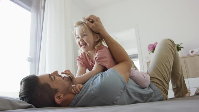 Happy young man lying on bed, holding his little daughter and having fun indoors at home.