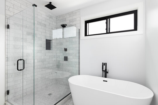 A renovated, luxury shower and freestanding bathtub with marble subway tiles, black faucet and shower head, and glass surrounding the shower.