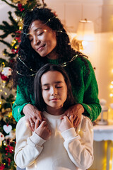 Brunette girl and long curly haired African American mother pose with illuminating garland on head and smile happily against Christmas tree