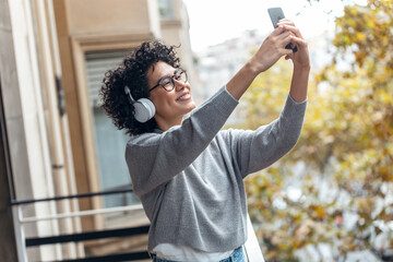 Beautiful woman taking photos with her smartphone while listening to music standing in the balcony at home.