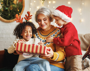 Two boys of different races giving New Year present to grandmother while sitting near xmas tree