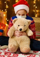 Child girl as Santa helper posing in new year decoration. Wearing a protective face mask against viruses, coronaviruses. She's playing with a toy bear. 