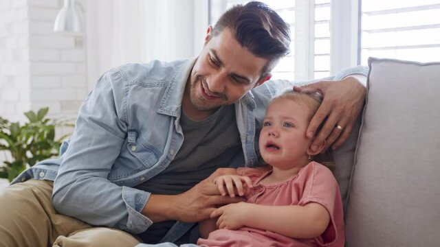 Young father sitting on sofa, holding and calming down his little daughter indoors at home.