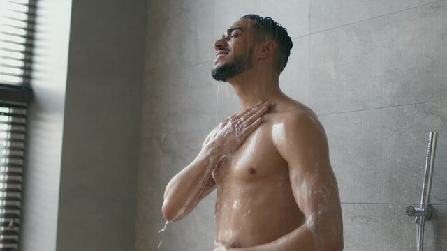 Young positive muscular middle eastern guy taking shower in morning, standing under water drops and washing his body