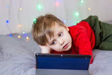 A cute boy is on the bed looking at a tablet. Online training, communication on the Internet. Funny smiling child.