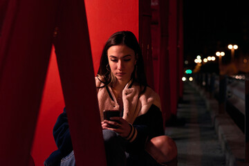 Young woman is sitting and is looking at her telephone on a red bridge