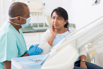 Fototapeta premium Latina suffering from toothache talking to stomatologist while sitting on dental chair in clinic
