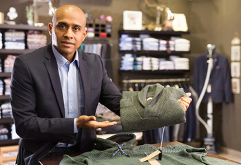 Portrait of young adult male seller suggesting sweater in clothing store