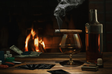Glass of cognac, cigar, bottle, cards game and chips on the table near the burning fireplace. Rest,...