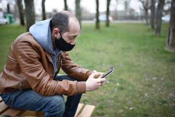 Caucasian man wearing a mask to protect himself from covid19 looks at his cell phone on a wooden park bench, open space.
