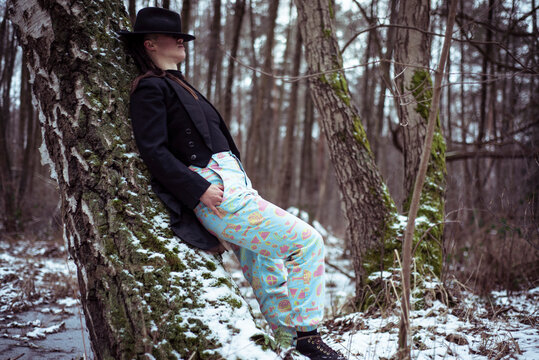 fashionable androgynous person with hat over eyes leans on snowy tree