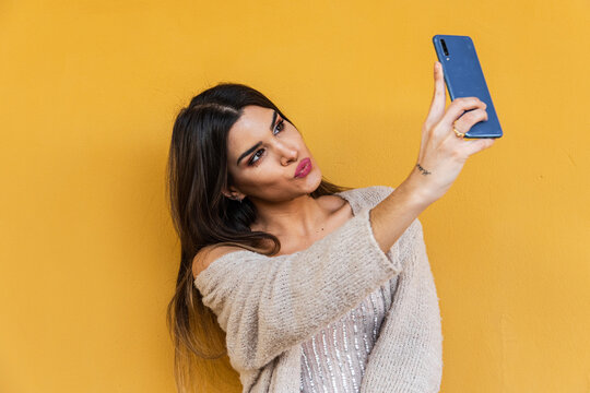 Portrait of spanish brunette girl taking a selfie with mobile phone on yellow background wall.