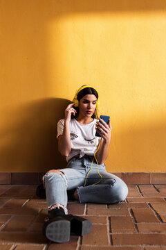 Portrait of Spanish brunette girl sitting on the floor and listening to music with headphones on yellow background wall.
