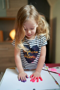 Cute little girl putting painted palms on white sheet of paper.